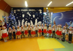 Grupa dzieci śpiewa kolędę, w tle dekoracja świąteczna, choinki.